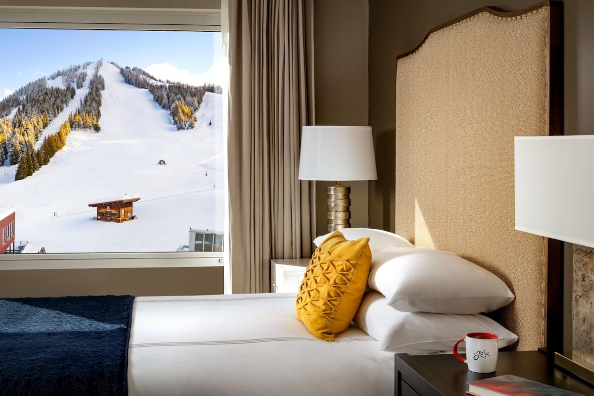 A hotel room with a bed, a nightstand, a lamp, a coffee mug, and a view of a snowy mountain landscape.
