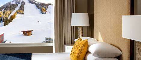 Une chambre d’hôtel avec un lit, une table de chevet, une lampe, une tasse à café et une vue sur un paysage montagneux enneigé.