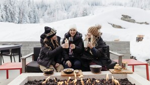 Tre personer sitter runt en eldstad och äter och dricker, i en snötäckt utomhusmiljö med berg i bakgrunden.