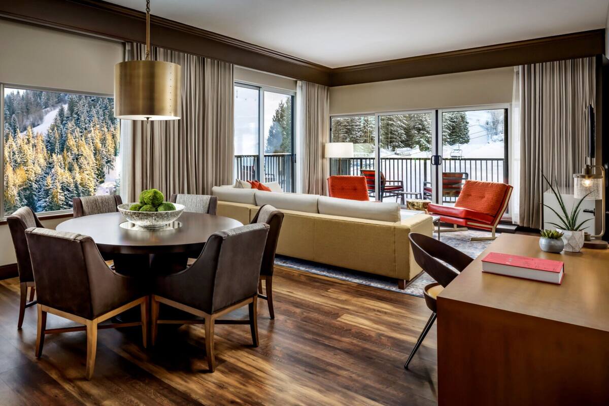 A modern hotel room with a dining area, a sofa, a balcony with outdoor seating, and a large window offering a view of trees.