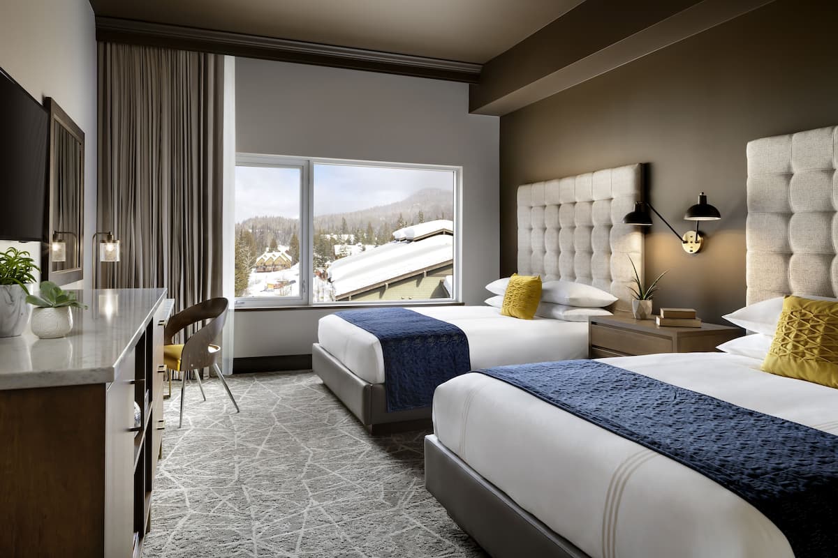 A modern hotel room with two beds, a desk, and a large window offering a view of a snowy landscape.