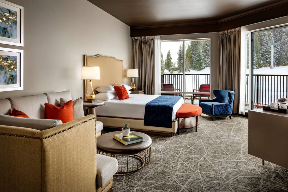 A hotel room with a large bed, a sofa, a chair, a small table, and a balcony with a view of trees.