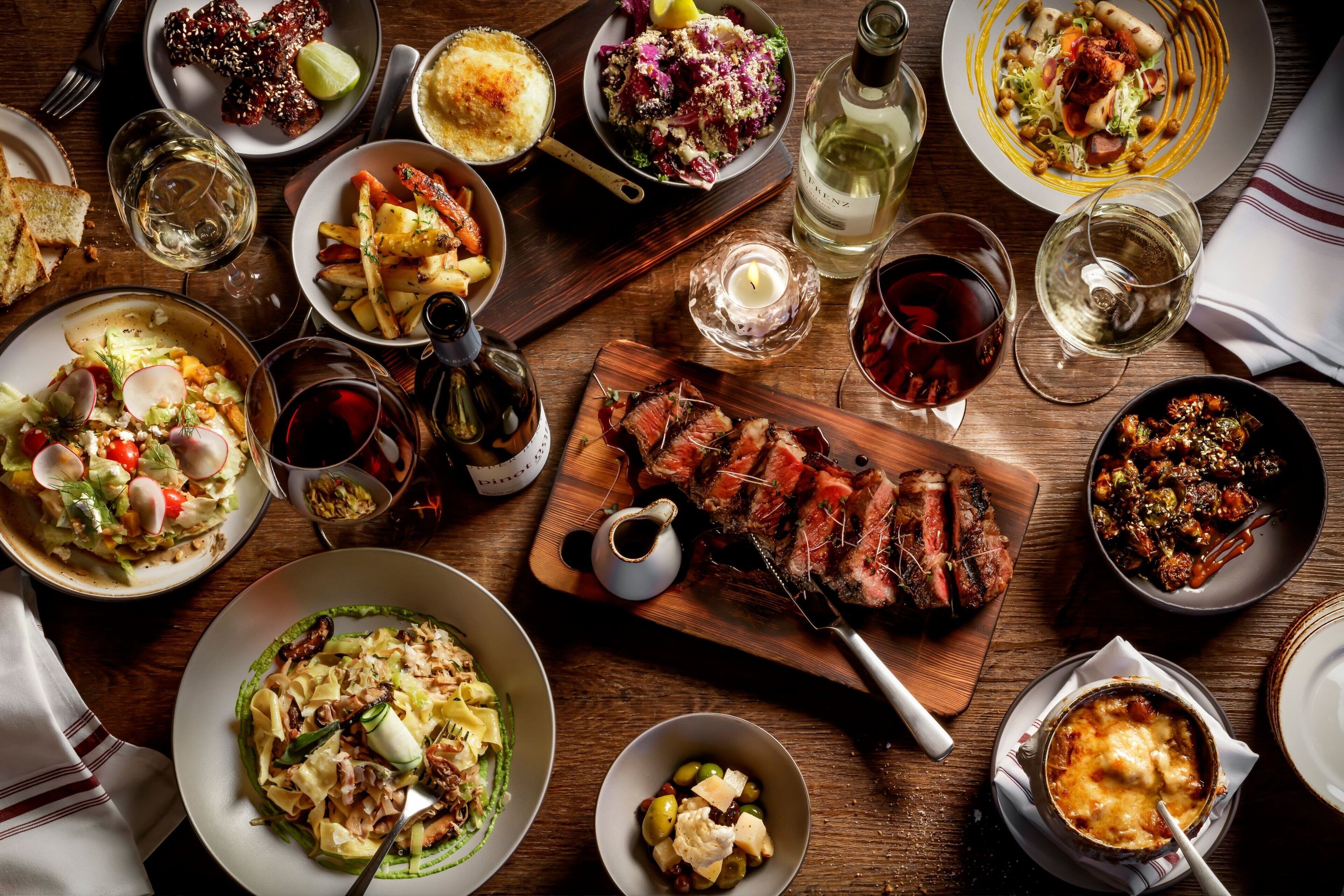 Variedad de platos y bebidas sobre una mesa de madera, incluyendo carne, ensaladas y recipientes con comida.