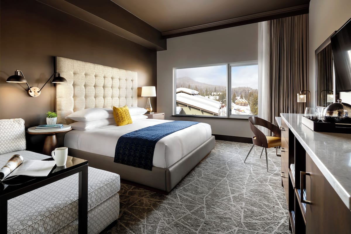 A hotel room with a large bed, a desk, a chair, a TV, and a window with a view of a snowy landscape.