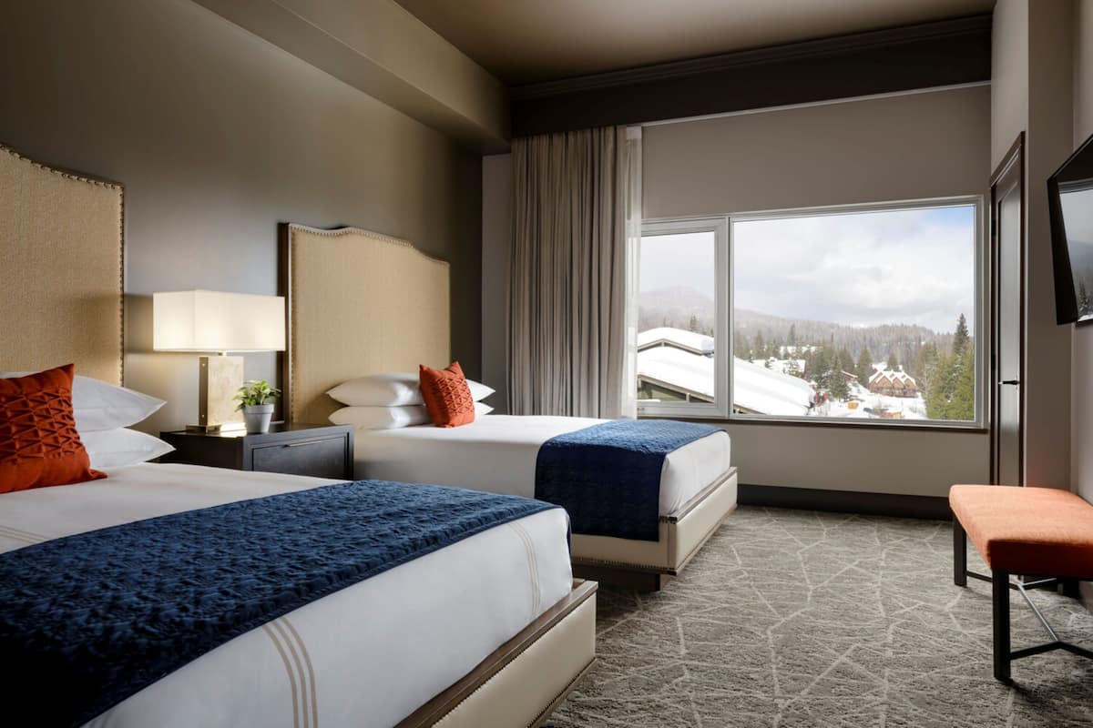 A hotel room with two beds, a large window with a view of a snowy landscape, a television mounted on the wall, and a bench.