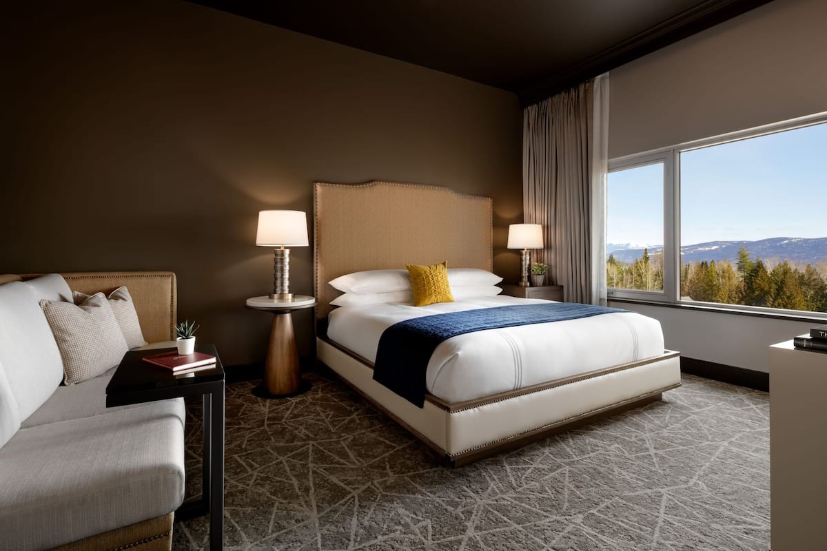 A hotel room with a large bed, a sofa, two bedside tables with lamps, and a view of the mountains through a large window.