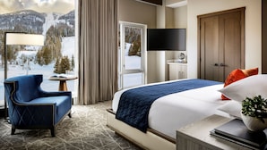 Ett hotellrum med en stor säng, en blå fåtölj, ett litet bord med en lampa, en tv och utsikt över snöklädda berg.