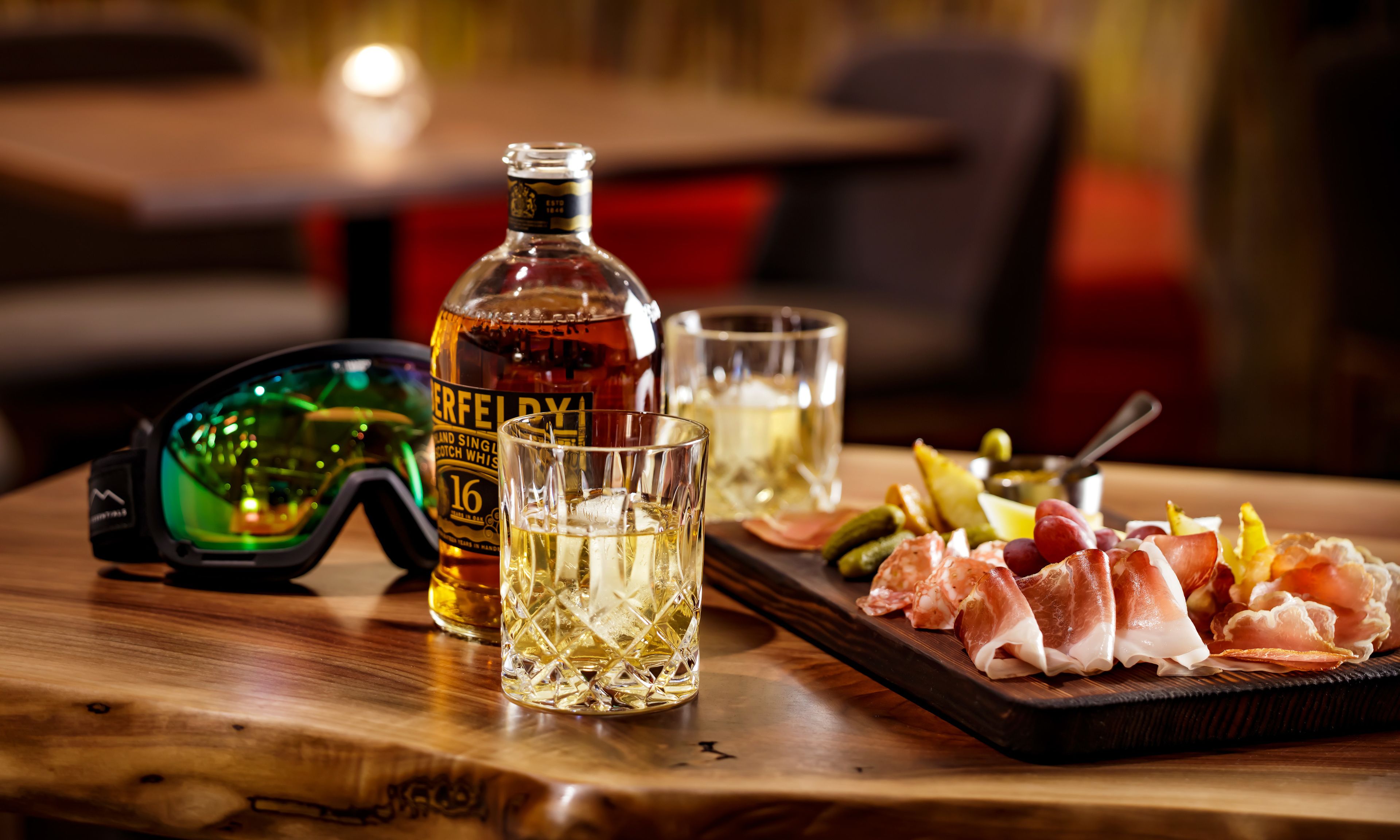 A bottle of whisky, two glasses, and a platter of meats on a wooden table.