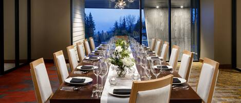 A long dining table set for a formal meal with chairs, a chandelier, and a view of trees through large windows.