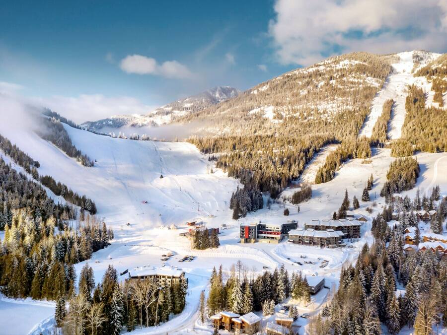 A snow-covered mountain resort with multiple buildings, including a lodge and a ski lift, surrounded by dense forests.