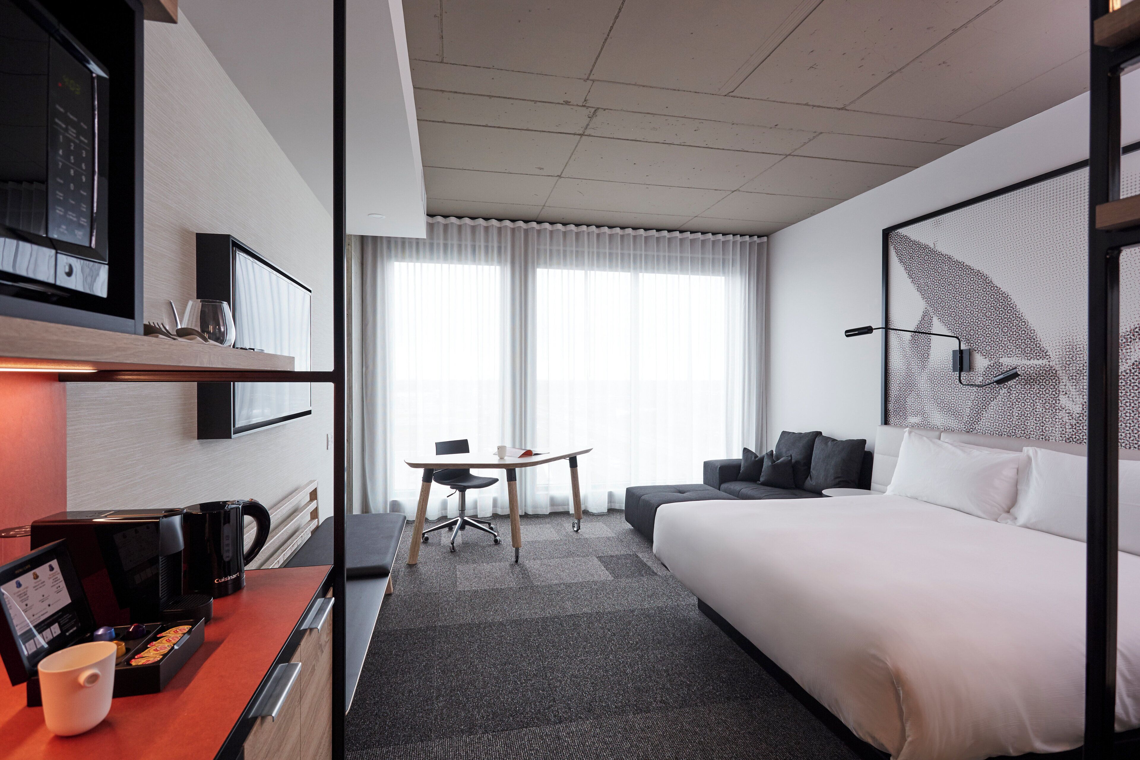 A modern hotel room with a large bed, a desk with a coffee maker, a TV, and a window with curtains.