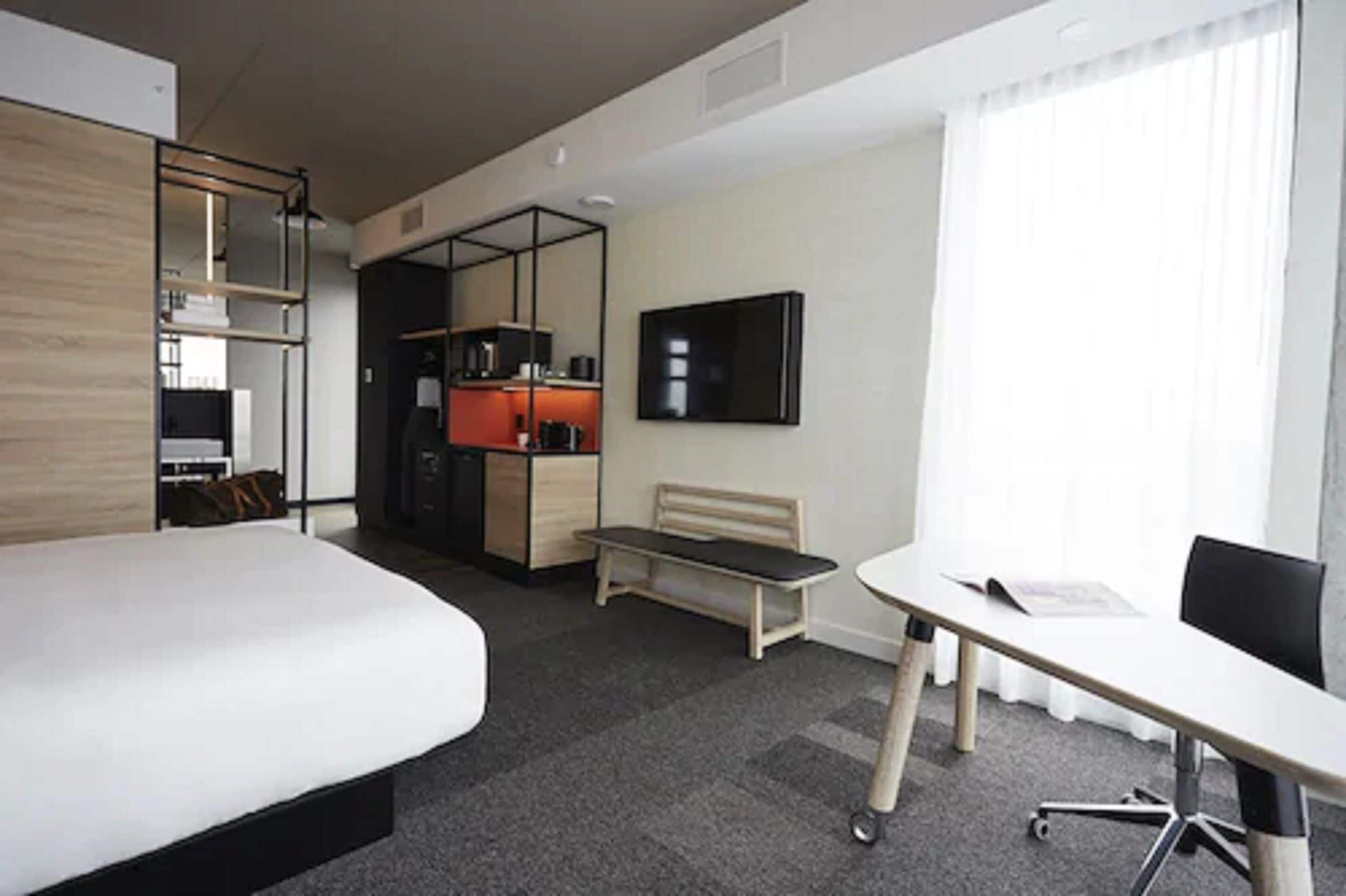 A hotel room with a bed, a desk, a chair, a television, and a kitchenette.