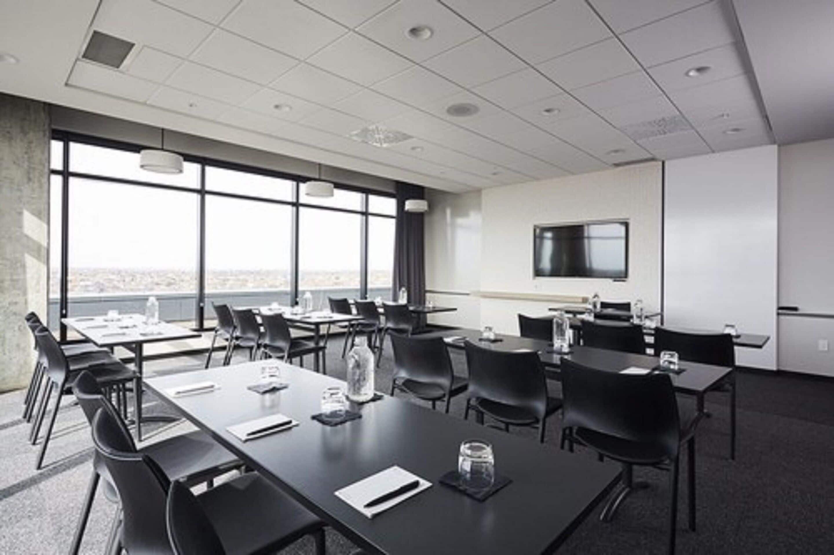 A conference room with tables and chairs, a large window with a city view, a flat-screen TV, and a kitchenette area.