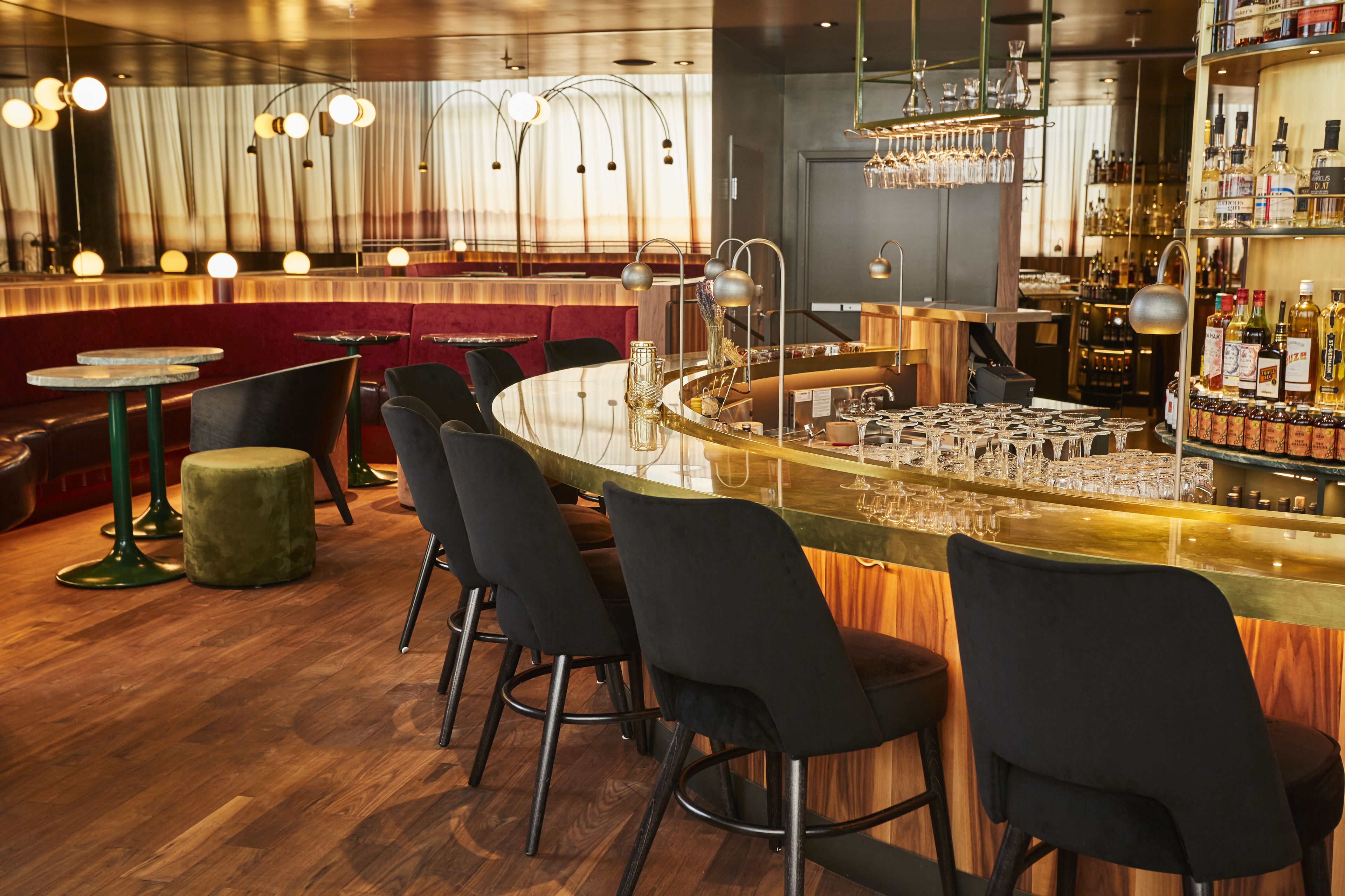 A bar area with a curved counter, multiple stools, and a variety of bottles and glasses on shelves.