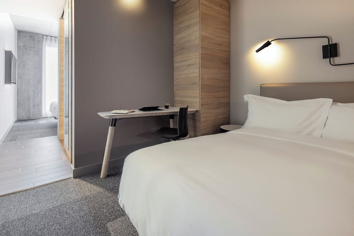 Ein modernes Hotelzimmer mit Bett, Schreibtisch, Stuhl, Nachttisch und Wandlampe.