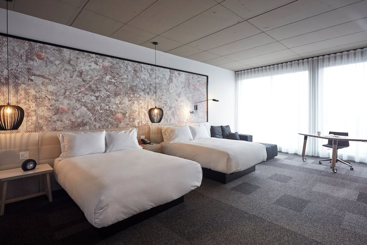 Ein Hotelzimmer mit zwei Betten, einem großen abstrakten Kunstwerk an der Wand, einem Schreibtisch mit Stuhl und einem großen Fenster mit Vorhängen.