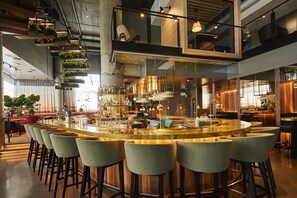 A modern bar area with a central bar, multiple stools, hanging plants, and a kitchen in the background. - Escad Hotel Quartier DIX30 (Brossard)