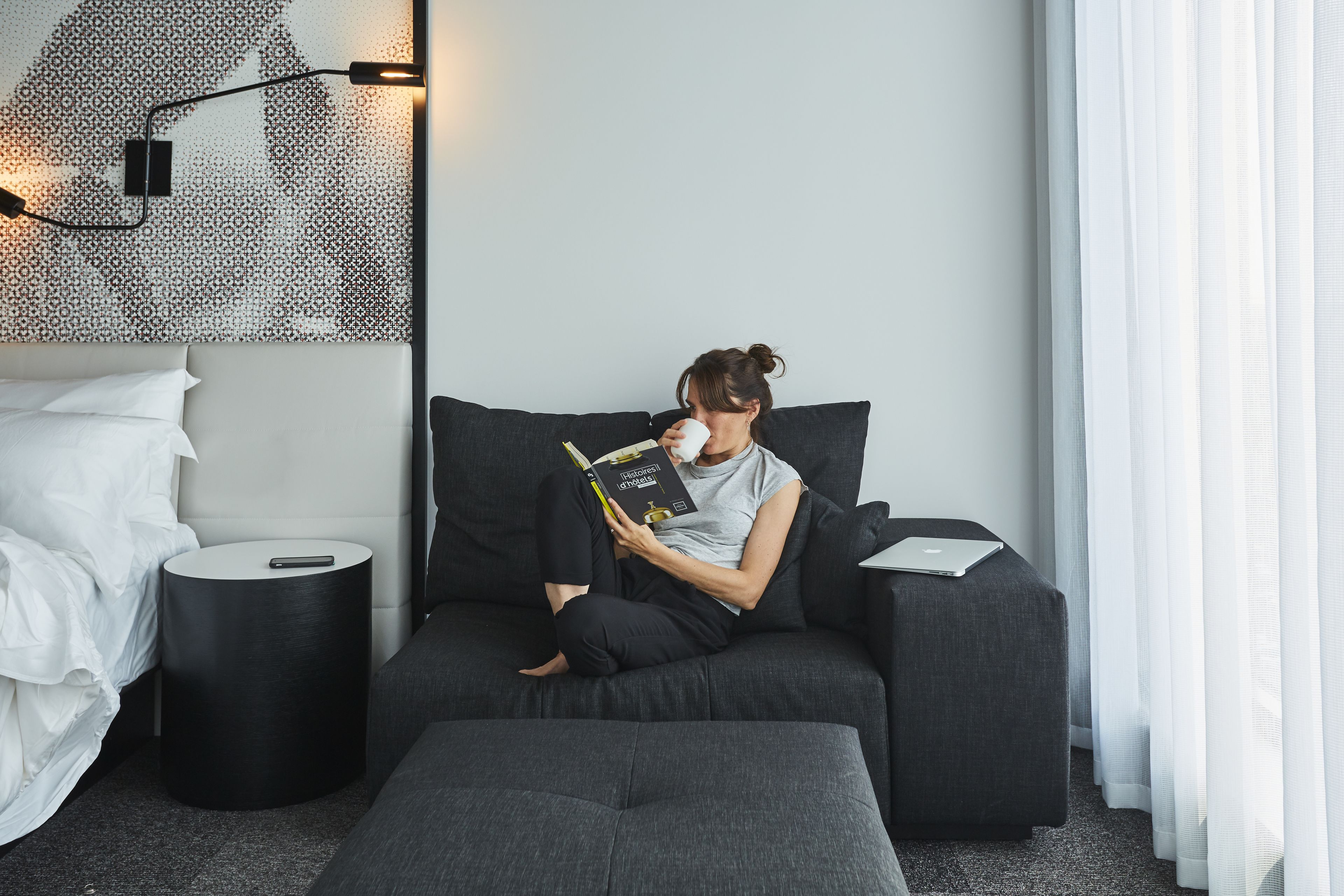 A modern room with a grey sofa, a small round side table, a white bed, and a patterned wall hanging.