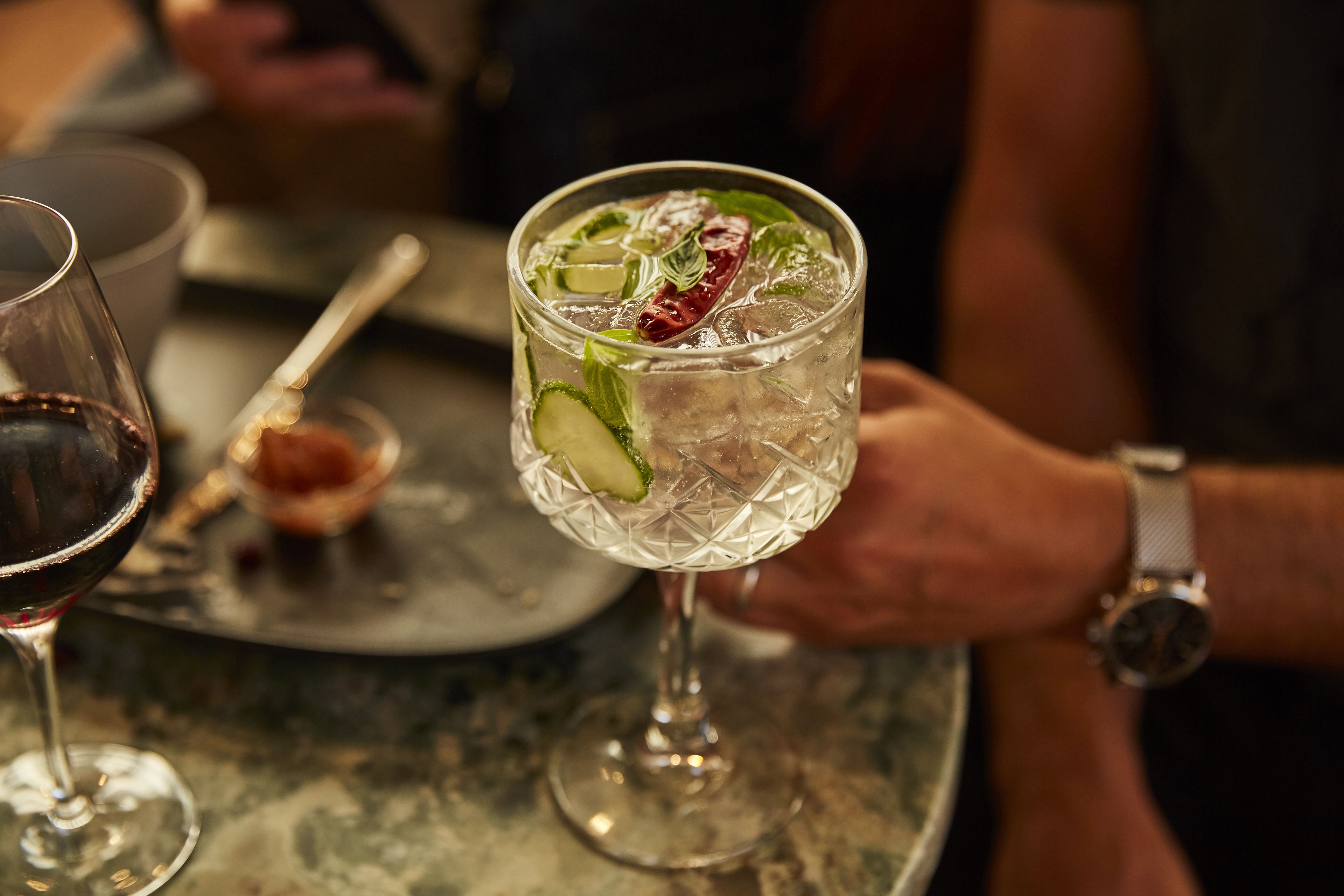 A glass of cocktail with ice and garnishes, held by a person wearing a watch.