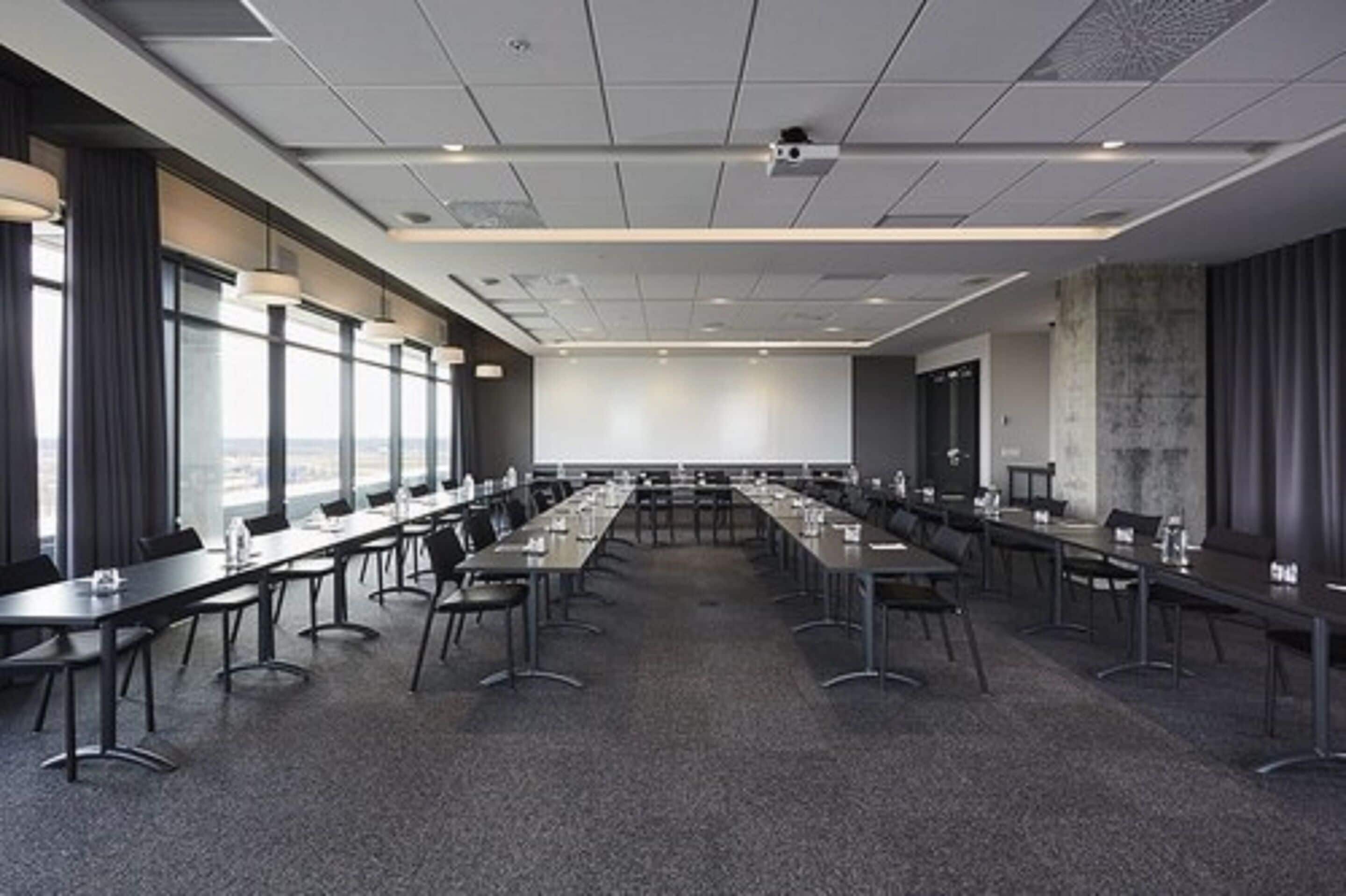 Uma sala de conferências com mesas compridas, cadeiras e uma tela grande na frente.