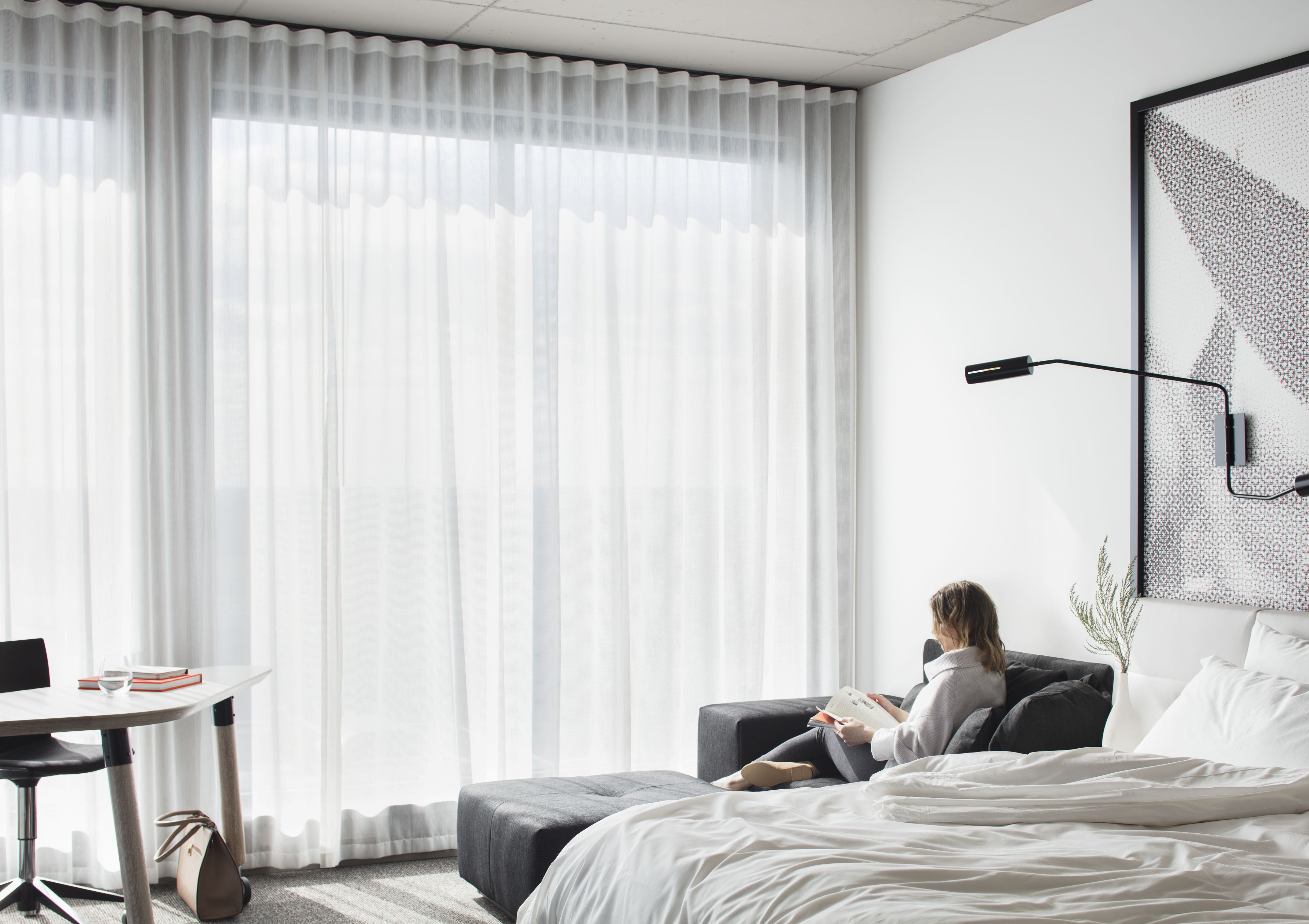 A modern hotel room with a large window, a bed, a reading chair, a desk, and a wall-mounted lamp.