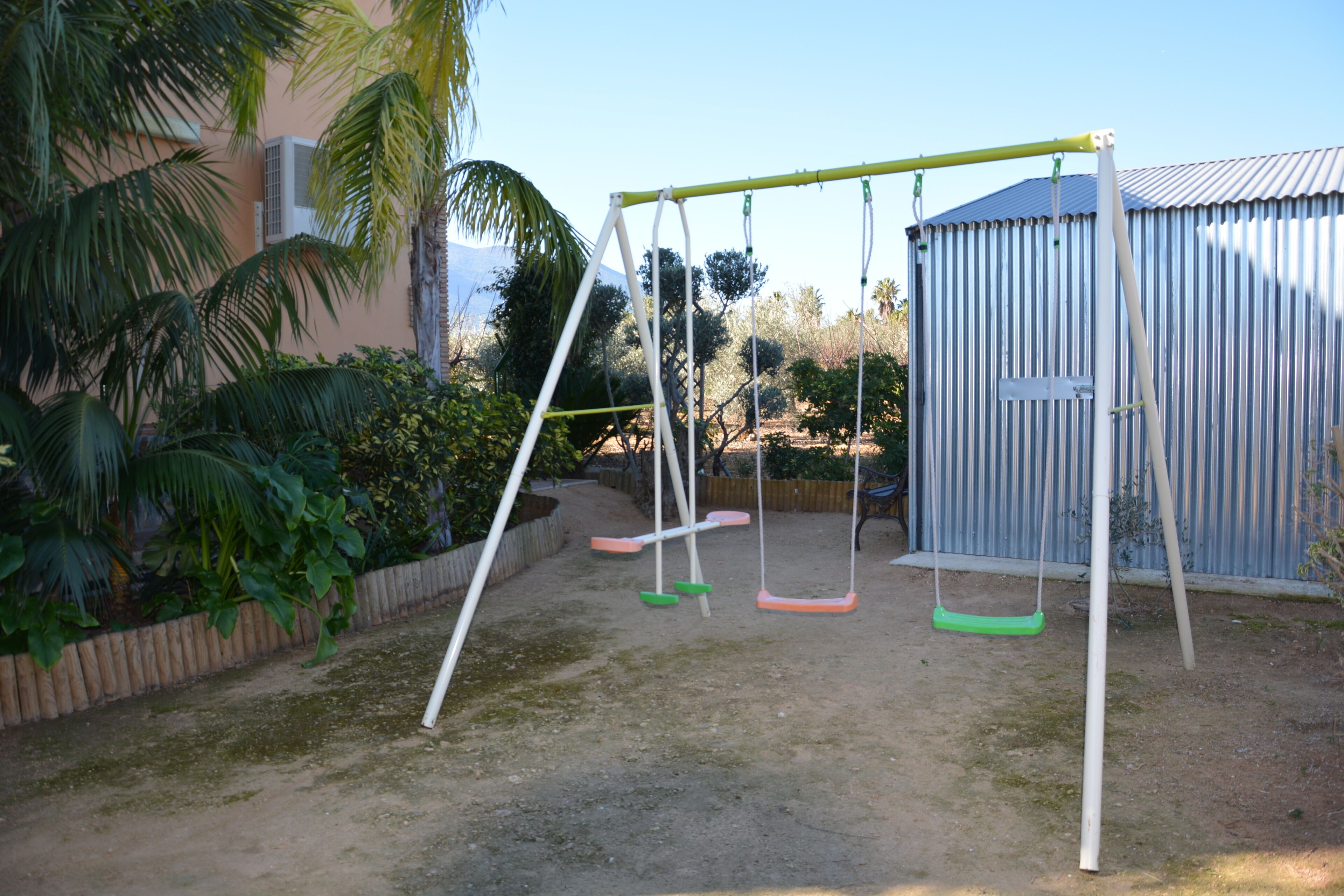 A swing set in a backyard with a wooden fence, a metal shed, and a palm tree.