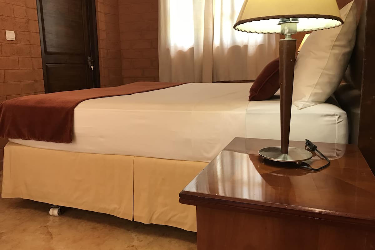 A hotel room with a bed, a bedside table with a lamp, a door, and a window with curtains.
