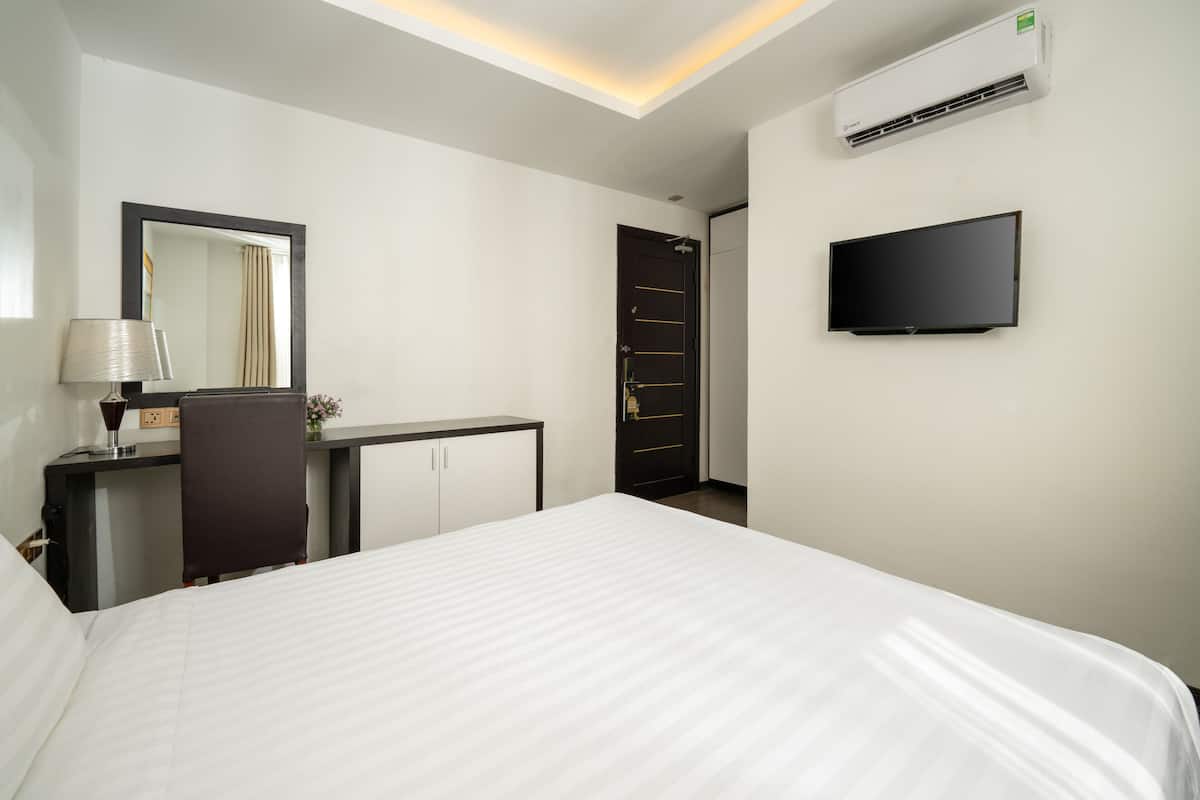A hotel room with a bed, a desk with a lamp, a wardrobe, a television, and an air conditioning unit.
