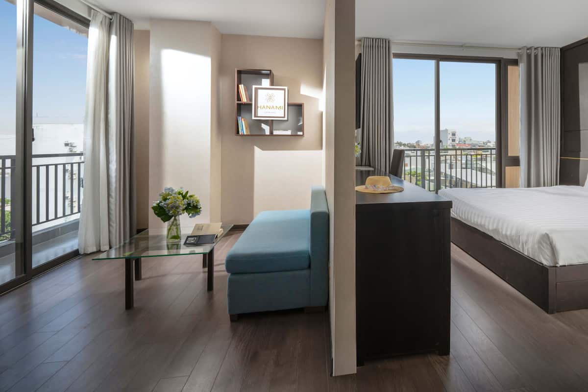 A modern hotel room with a large bed, a glass coffee table, a blue armchair, and a view of the city.