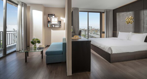 A modern hotel room with a large bed, a glass coffee table, a blue armchair, and a view of the city. - Hanami Hotel Danang (Da Nang)