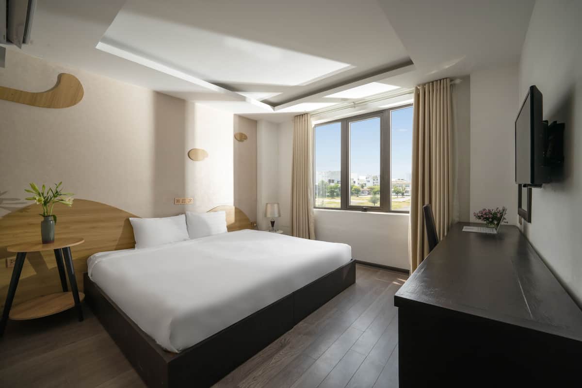 A modern hotel room with a large bed, a desk, a TV, and a window with a city view.