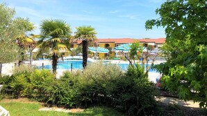 A pool area with palm trees, umbrellas, and a building with a red roof. - Enjoy the sun from a private garden! (San Michele Al Tagliamento)