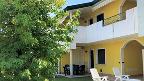 A two-story building with a yellow facade, a balcony with white railings, and a patio area with outdoor furniture. - Enjoy the sun from a private garden! (San Michele Al Tagliamento)