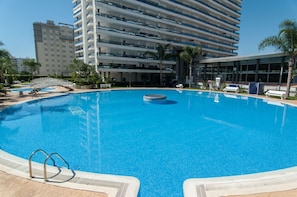 Una piscina grande y cristalina con una fuente central, rodeada por una zona de piscina con sillones de descanso y una palmera.