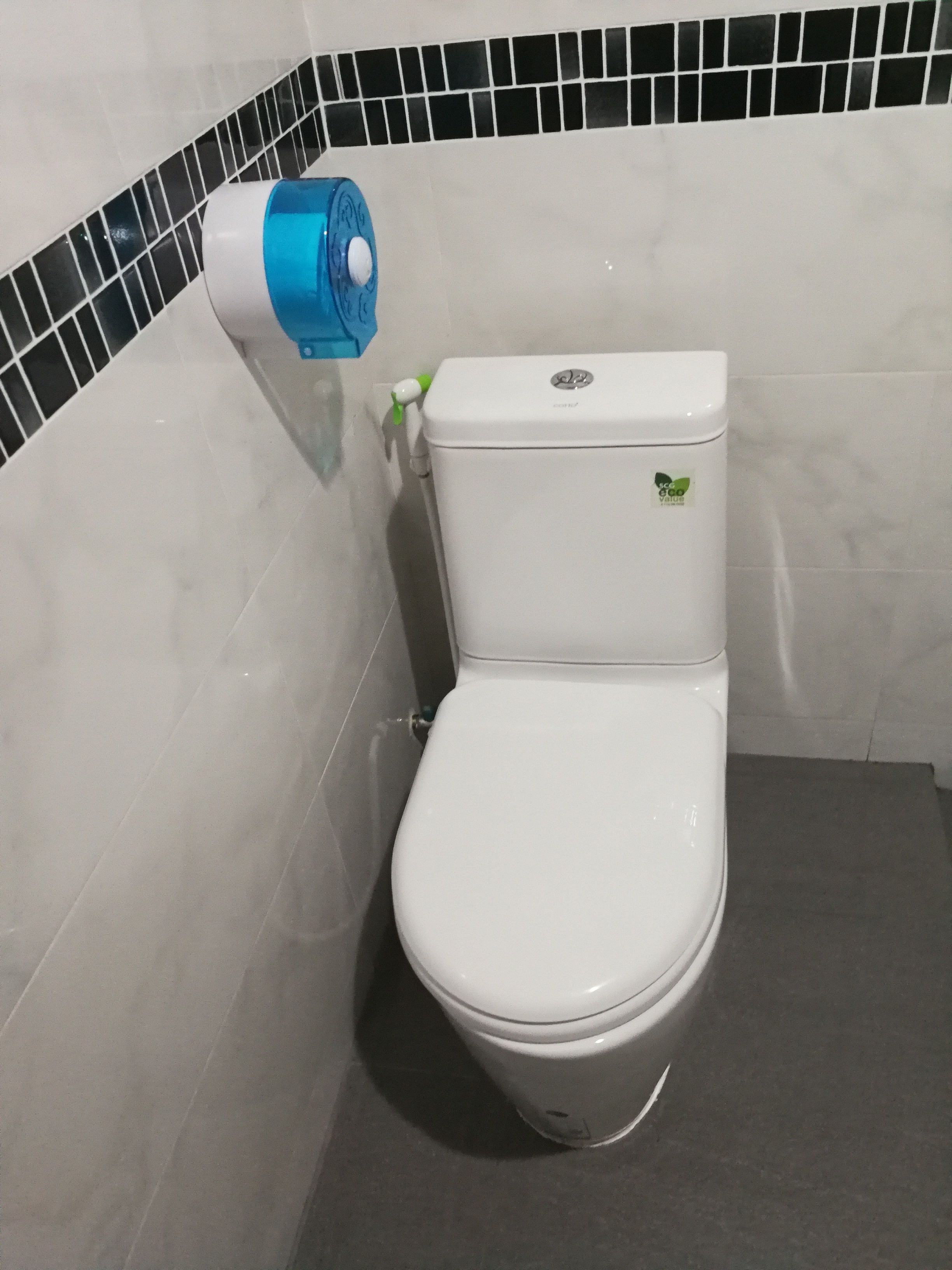 A white toilet with a blue bidet attachment, a green sticker on the tank, and a black and white tiled backsplash.