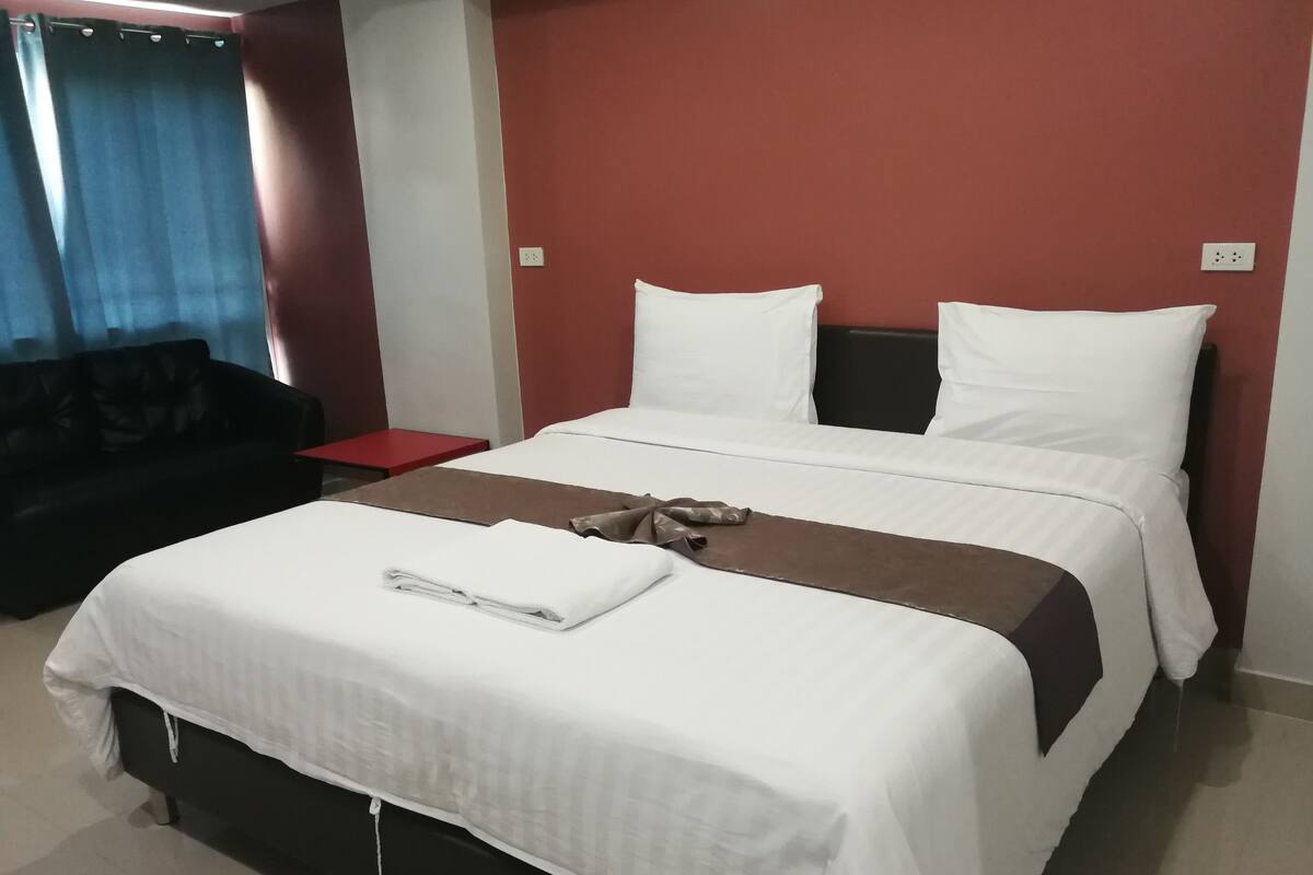 A neatly made bed with white and brown bedding, a red nightstand, and a dark-colored sofa in a room with red walls.