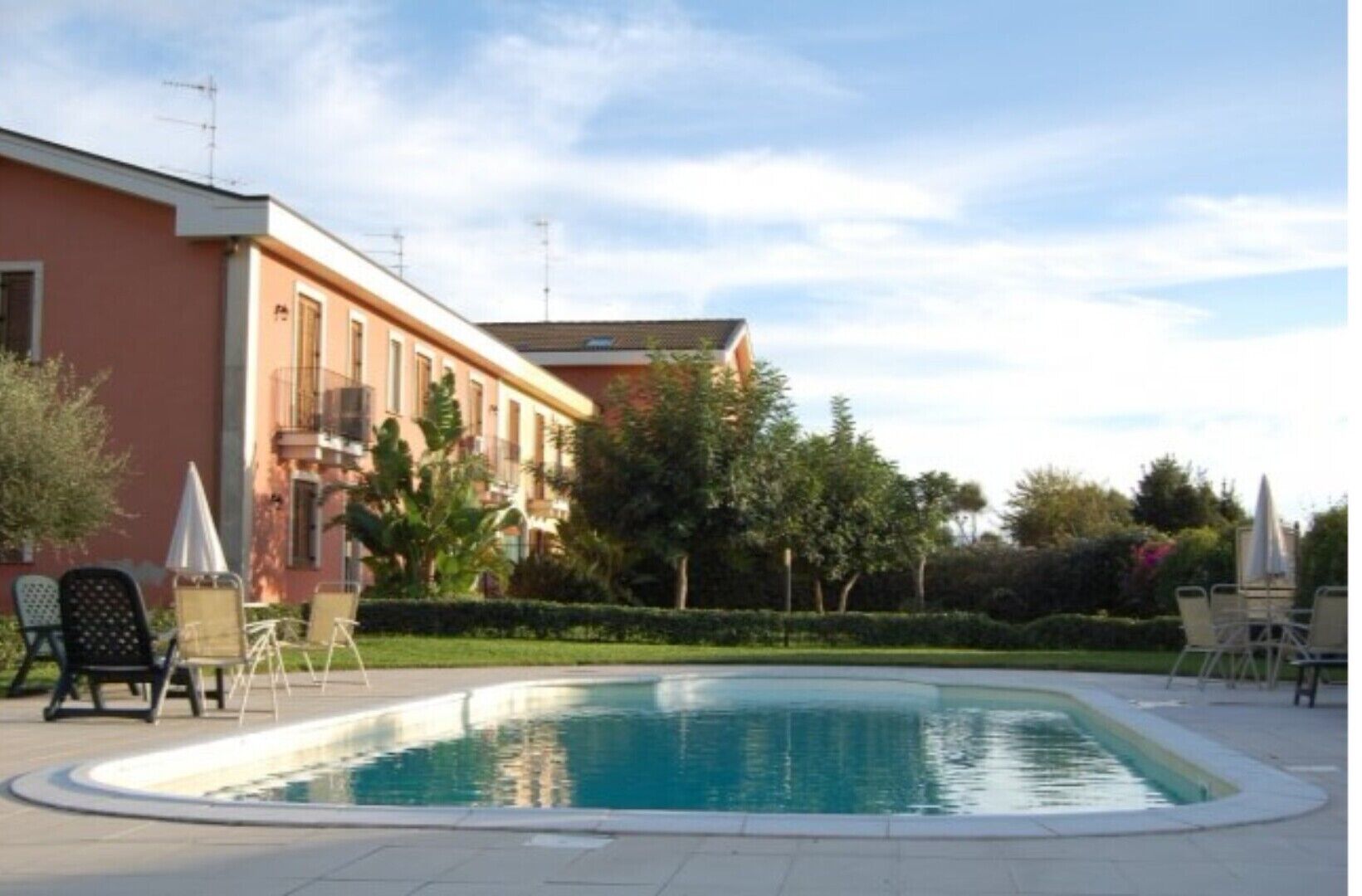 A swimming pool with lounge chairs and umbrellas, surrounded by a landscaped garden and a building with balconies.