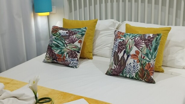 Quarto de hotel com cama, almofadas, mesa de cabeceira, abajur e pintura de uma paisagem costeira.
