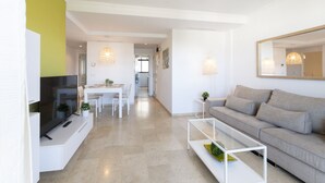 Ein modernes Wohnzimmer mit Flachbildfernseher, grauem Sofa, Glastisch und Essbereich mit weißem Tisch und Stühlen.