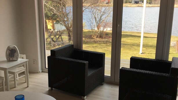 Ein Zimmer mit einer großen Schiebeglaswand, die auf einen See Blick gewährt, zwei schwarze Rattan-Sofas, ein weißer Tisch mit einer blauen Kerze und ein Ventilator auf einem kleinen Tisch.