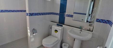 Un baño con inodoro, lavamanos y espejo. Presenta un rebozado de azulejos azules y blancos, un dispensador de papel higiénico blanco y un bote de basura blanco.