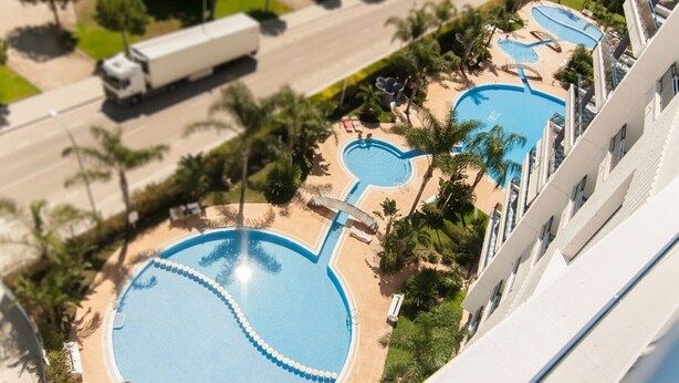 Un hotel con varias piscinas, palmeras y una carretera con vehículos.