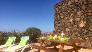 A wooden table set with a green placemat, a glass of orange juice, and a bowl of fruit. There are two lounge chairs with green cushions. - ECO Friendly Volcanic Rock Villa with Private Pool (6.7m x 4.6m) WiFi. Sea View. (Villa Verde)