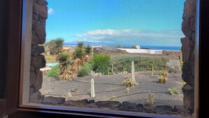 A view through a window with stone frames, featuring a garden with cacti and a distant landscape with mountains and a clear sky. - ECO Friendly Volcanic Rock Villa with Private Pool (6.7m x 4.6m) WiFi. Sea View. (Villa Verde)