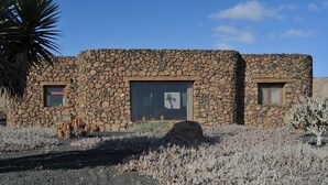 A stone building with two windows and a large door, surrounded by desert vegetation. - ECO Friendly Volcanic Rock Villa with Private Pool (6.7m x 4.6m) WiFi. Sea View. (Villa Verde)