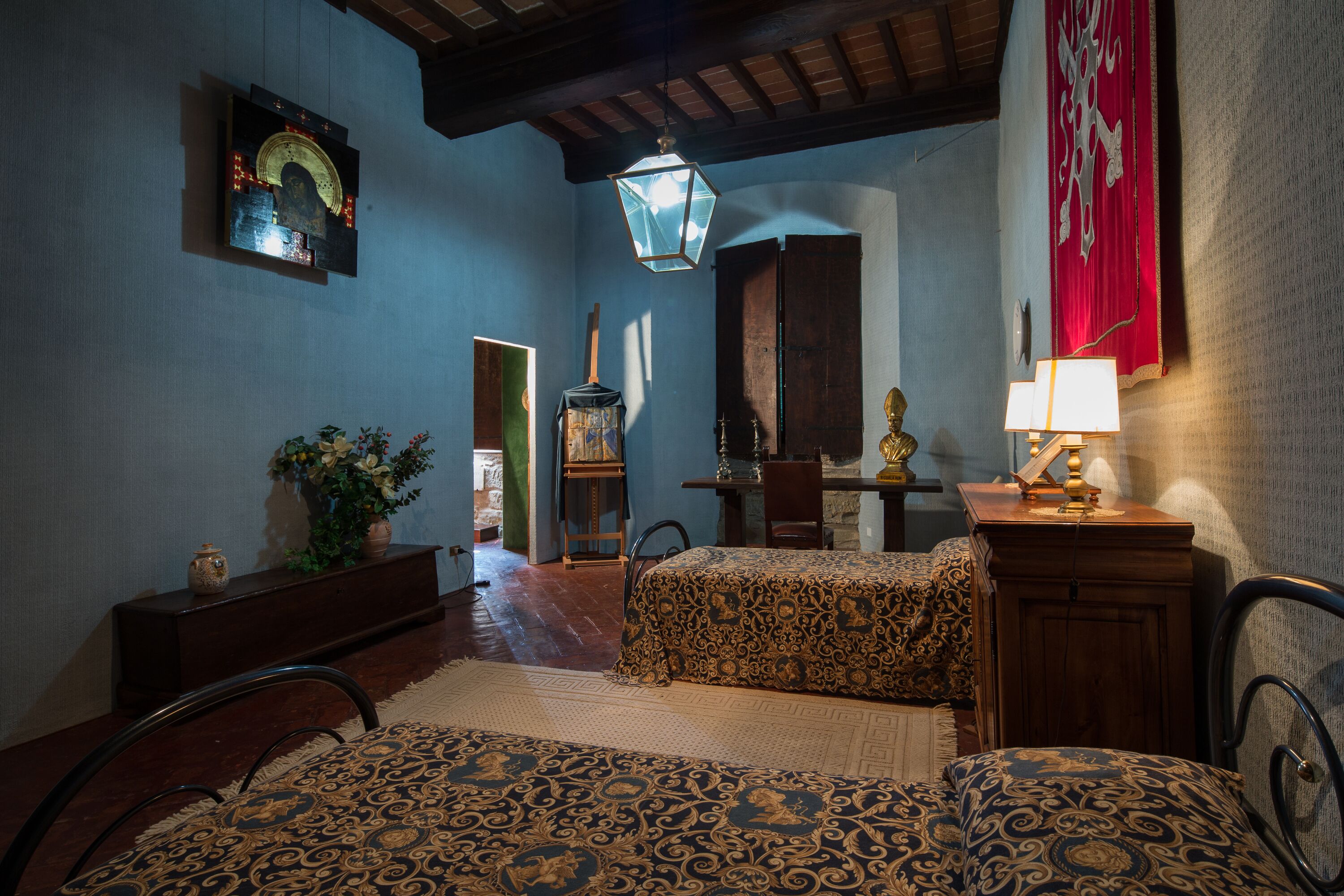 A traditional room with a bed, a desk, and a chair. There is a lamp on the desk and a plant on the dresser.