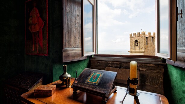 A room with a view of a castle through a window, a wooden table with a book and candle, and a painting on the wall. - 3BR luxury apt, walk to downtown Cortona, and amazing view over the town hall (Cortona)