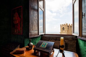A room with a view of a castle through a window, a wooden table with a book and candle, and a painting on the wall. - 3BR luxury apt, walk to downtown Cortona, and amazing view over the town hall (Cortona)