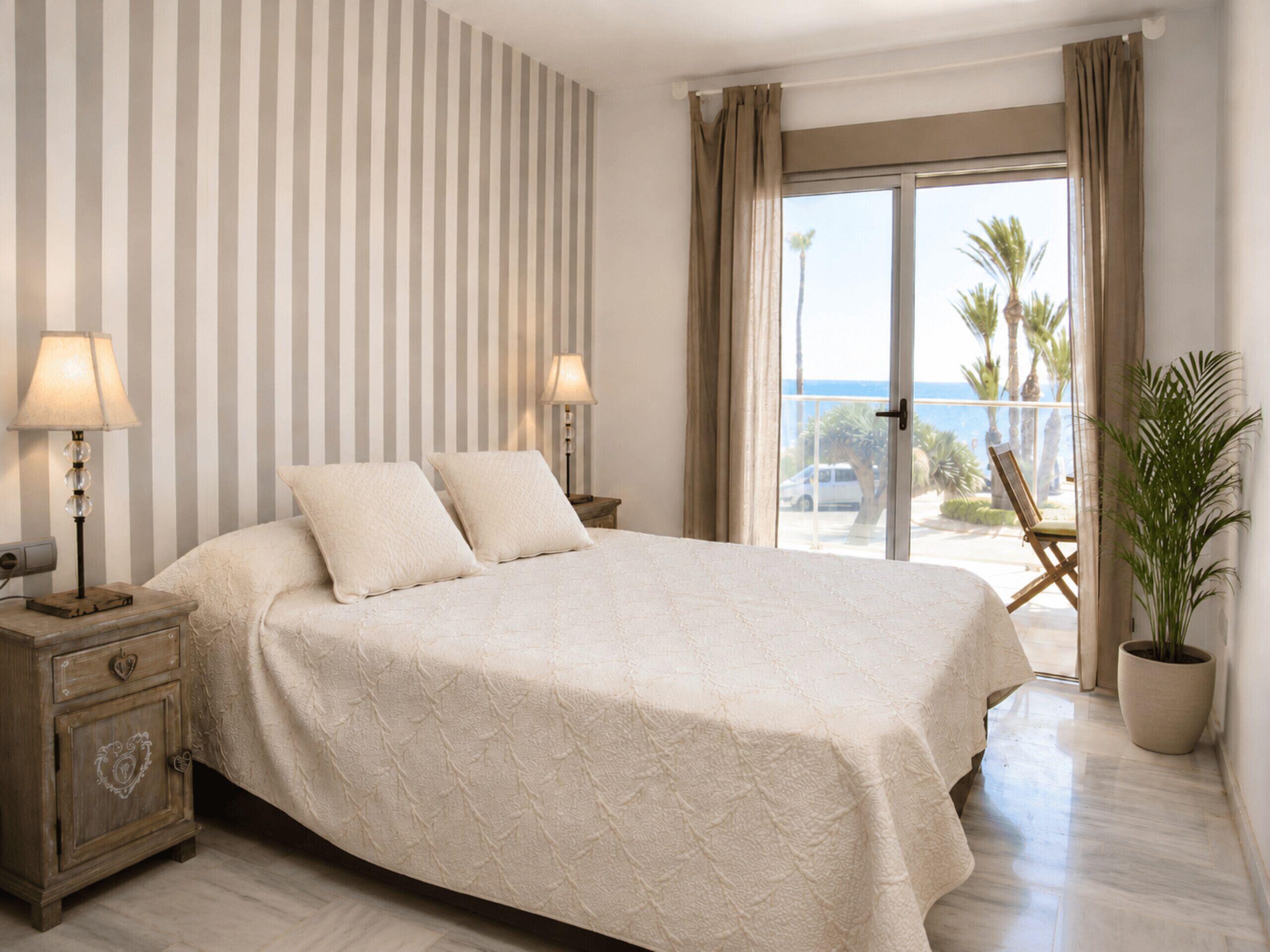 Habitación de hotel con una cama grande, dos lámparas de noche, una mesita de noche de madera, una planta en maceta y vistas al mar a través de una puerta corrediza de cristal.