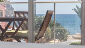 Blick aus einem Fenster auf einen Strand mit Segelboot, Palmen und einer felsigen Küste.